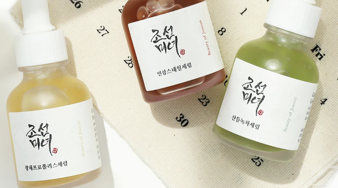 Tre flaskor från koreanska hudvårdsmärket Beauty of Joseon placerade på en ljus tygbakgrund med tryckt kalenderdesign. Flaskorna innehåller olika serum i gul, brun och grön nyans, och etiketterna är tryckta med traditionell koreansk kalligrafi.