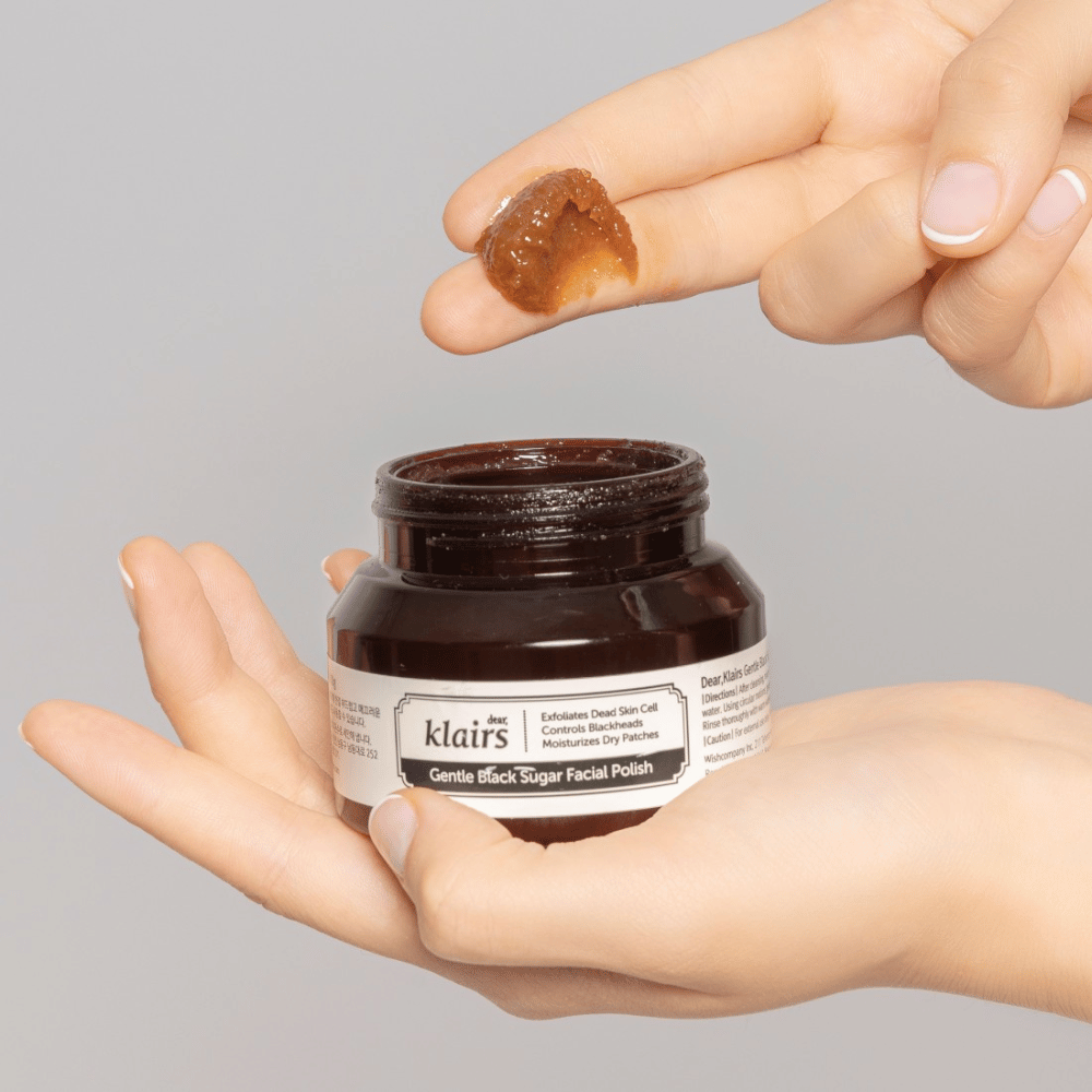 Händer håller en öppen burk av Klairs Gentle Black Sugar Facial Polish och visar produkten på ett finger, mot en grå bakgrund.